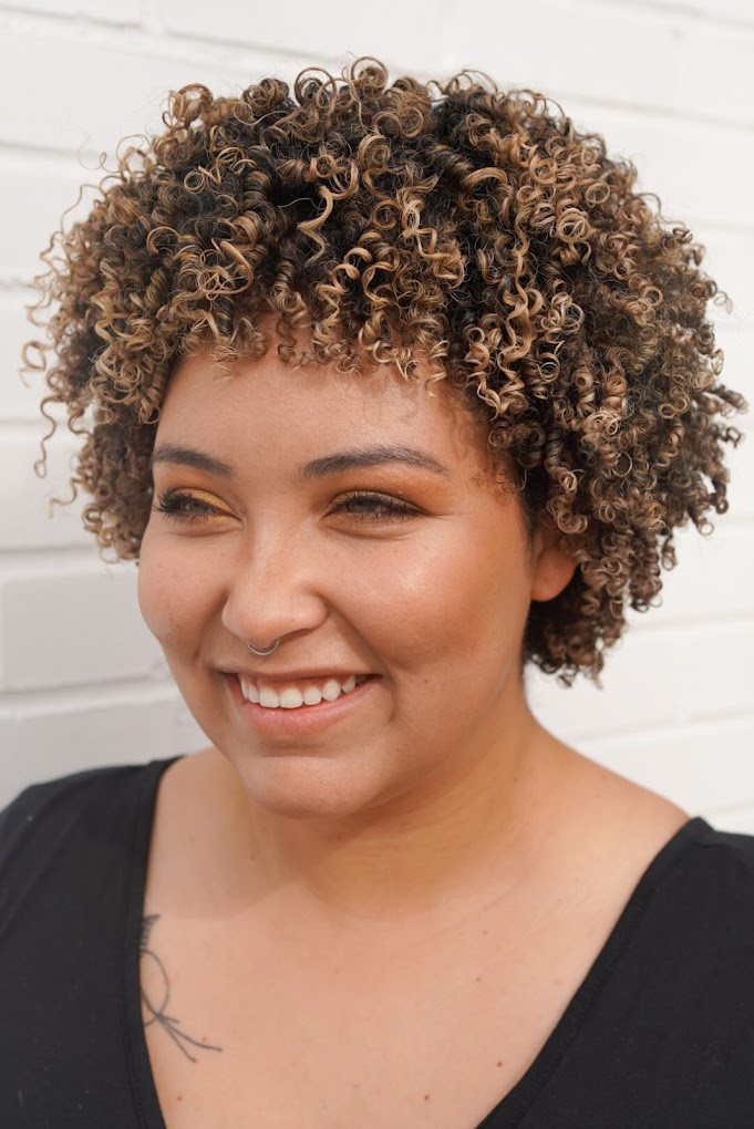 Smiling guest with short, springy coils with golden highlights against a white brick wall