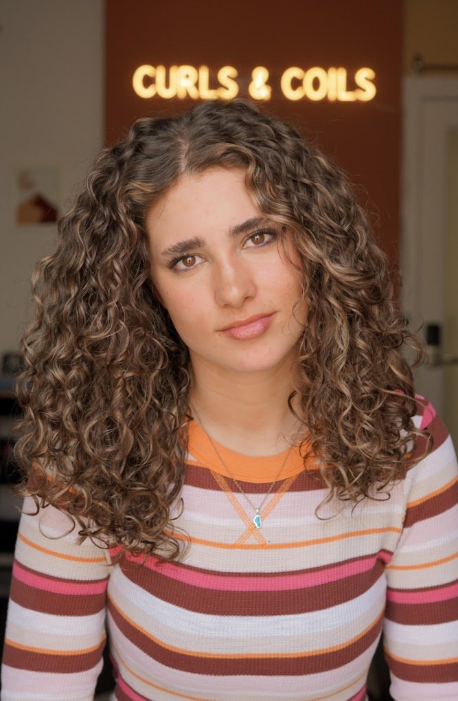 Guest with long, defined brown curls with caramel highlights in front of the Curls & Coils sign